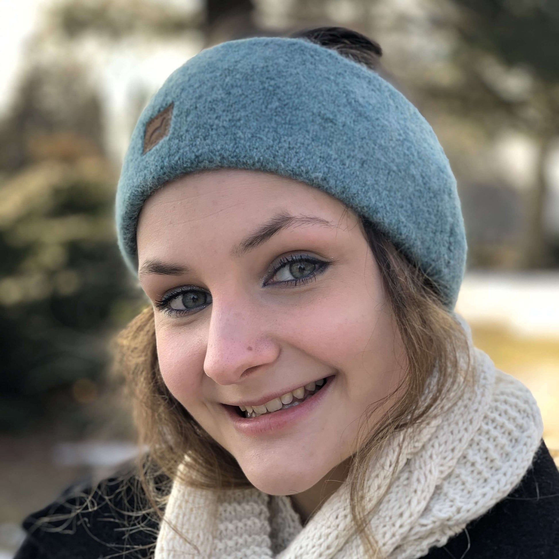 Woman wearing a light aqua green alpaca knit headband and an ivory knit scarf with a blurred outdoor background.