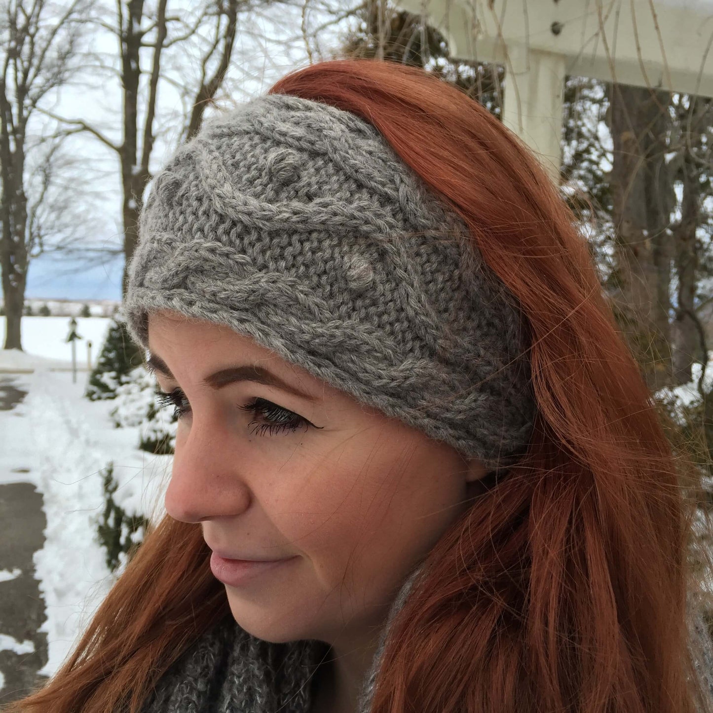 Side profile of woman wearing a cable knit alpaca headband in grey with winter background.