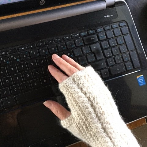 A close-up of a person's hand wearing a cabled cream-colored knit fingerless glove, resting on the keyboard of a black laptop.
