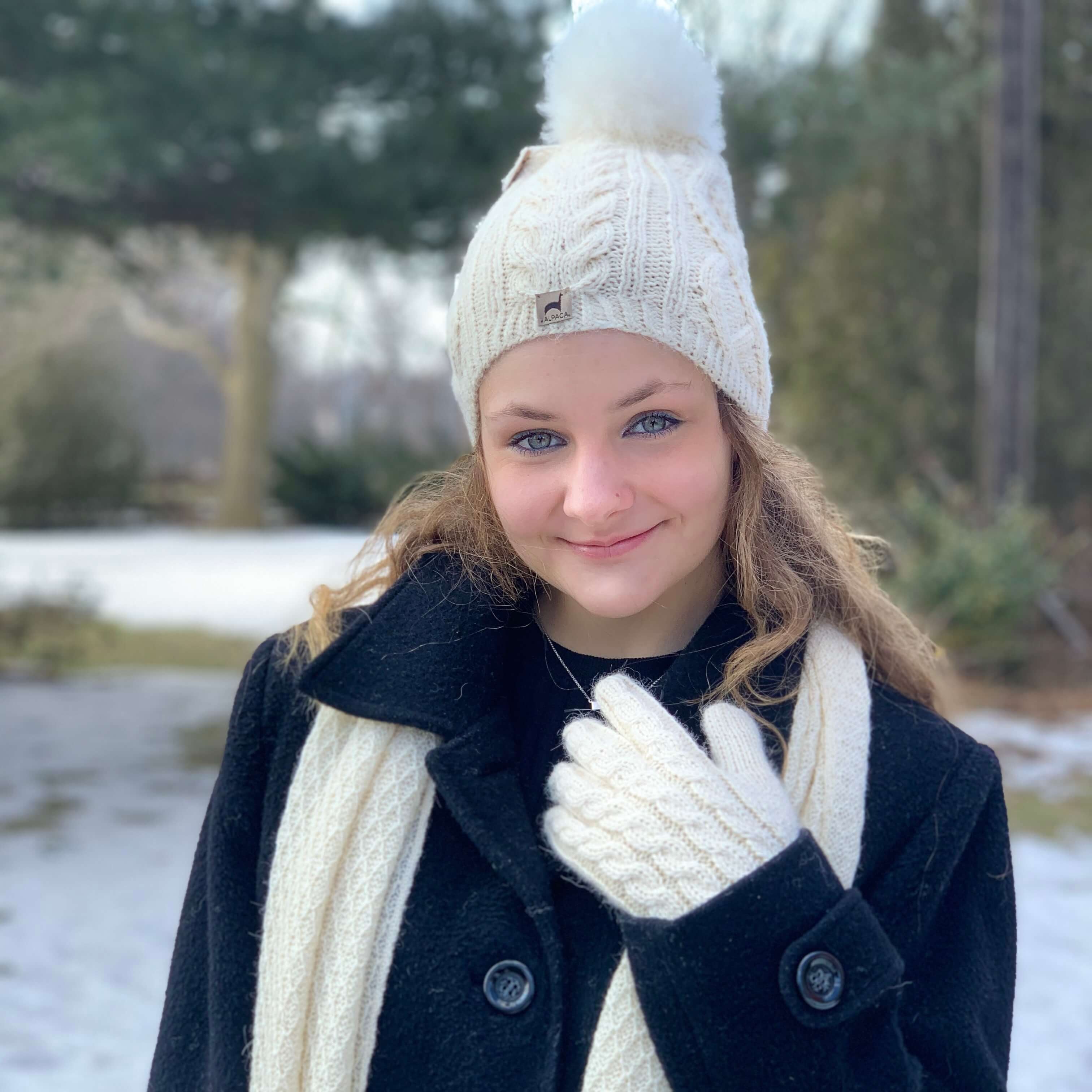 Person wearing a black coat, white alpaca scarf, and knit alpaca hat with a pom-pom and alpaca gloves in a snowy outdoor setting.